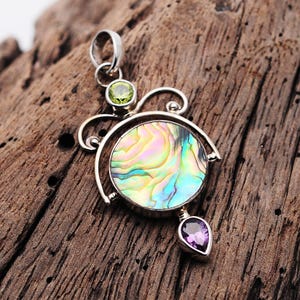May include: A silver pendant with a round abalone shell inlay, a green gemstone, and a purple gemstone. The pendant is on a wooden surface.
