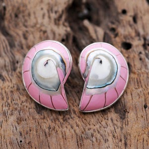 May include: A pair of pink and silver seashell-shaped earrings. The earrings have a spiral design with a pink enamel border and a pearlescent center. The earrings are set against a wooden background.