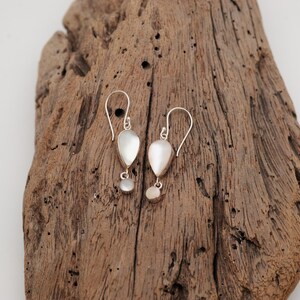 Könnte beinhalten: Ein Paar silberne Ohrhänger mit einer tropfenförmigen weißen Muscheleinlage und einem kleinen runden weißen Muschelakzent, die an einem gebogenen Silberdraht haken hängen. Die Ohrringe haben eine polierte metallische Oberfläche.