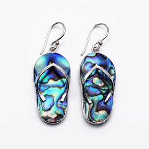 Op de afbeelding: Een paar zilveren oorbellen in de vorm van slippers. De oorbellen zijn gemaakt met iriserende abalone schelpen, met blauwe, groene en paarse tinten. Het zilver omlijst de schelp en de haak heeft een eenvoudig, gebogen ontwerp.