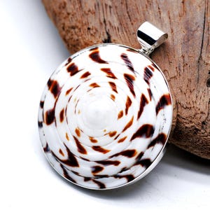 May include: A round pendant featuring a white seashell with brown spots, set in a silver-colored metal frame. The pendant has a loop at the top for a chain. The background is a piece of wood.