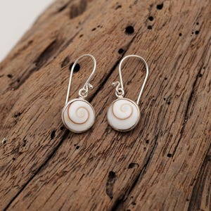 May include: A pair of silver drop earrings featuring circular shiva shell inlays. Each earring has a polished silver setting that holds a white shell with a natural brown spiral pattern. The earrings are suspended from simple silver ear wires.