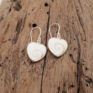 Peut inclure: Une paire de boucles d'oreilles en forme de cœur avec une incrustation de coquillage blanc présentant un motif en spirale naturel, serties dans une monture en métal argenté poli avec des crochets incurvés pour une fixation sûre.