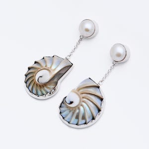Puede incluir: Un par de pendientes de plata con un diseño de concha de nautilus con colores iridiscentes y un acento de perla. Los colgantes en forma de concha están conectados a un perno de perla mediante una delicada cadena.