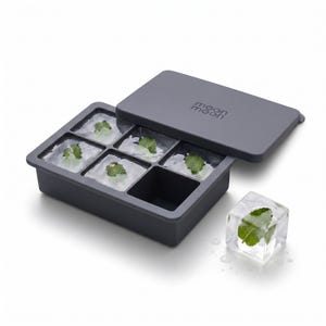 May include: A dark grey silicone ice cube tray with a matching lid featuring the text moon moon. The tray has six square compartments, five of which contain clear ice cubes with green mint leaves frozen inside, and one empty square slot.