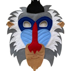 May include: A felt mask in the shape of a mandrill's face. The mask features a blue and red nose, blue cheeks, and a tan beard. The eyes are open, and the mask has gray, white, and black accents.