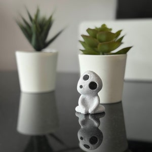 Puede incluir: Una pequeña figurita blanca impresa en 3D de un personaje de la película 'Mi vecino Totoro'. La figurita tiene una cabeza redonda con dos ojos negros y está sentada con las piernas cruzadas.