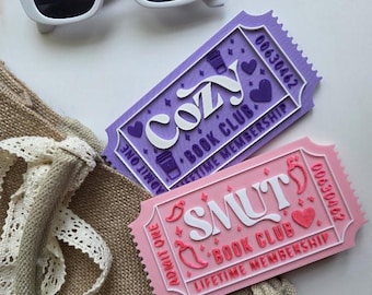 Puede incluir: Gafas de sol blancas sobre un bolso de mano de arpillera con ribete de encaje. Se ven dos membresías de club de lectura con forma de boleto: una morada con "Cozy" y otra rosa con "Smut". Ambos boletos tienen el texto "Book Club" y "Lifetime Membership".