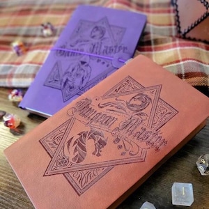 May include: Two leather journals with "Dungeon Master" embossed on the covers. One is reddish-brown, the other purple. The journals feature geometric designs and fantasy illustrations. A purple elastic band secures the purple journal. Dice are scattered around.