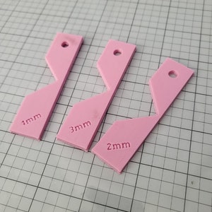 Op de afbeelding: Drie roze plastic stukken met verschillende openingen. De stukken zijn gelabeld met "1 mm", "2 mm" en "3 mm".