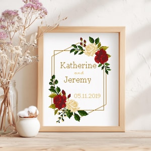 May include: A framed cross-stitch embroidery featuring a gold geometric border decorated with red and cream roses and green leaves. The text reads Katherine and Jeremy with the date 05.11.2019 in a gold-colored font on a white fabric background.