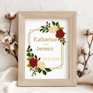 May include: A framed cross-stitch pattern featuring a gold geometric border decorated with red and cream roses and green leaves. The text reads Katherine and Jeremy with the date 05.11.2019 in the center. The design is displayed in a light wood frame.