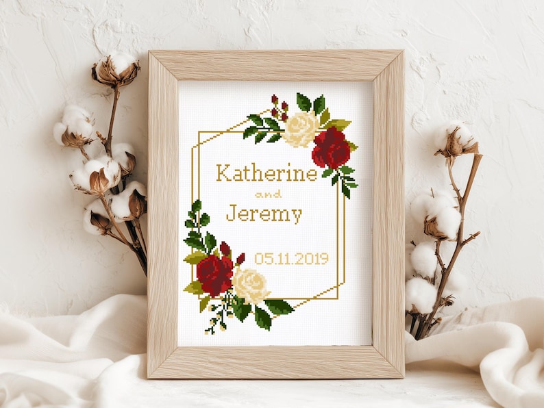 May include: A framed cross-stitch pattern featuring a gold geometric border decorated with red and cream roses and green leaves. The text reads Katherine and Jeremy with the date 05.11.2019 in the center. The design is displayed in a light wood frame.