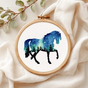 May include: A cross-stitch embroidery of a horse silhouette filled with a blue and green night sky pattern featuring stars and a forest of black pine trees, secured within a round wooden embroidery hoop frame.