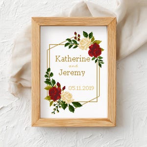 May include: A framed cross-stitch embroidery featuring a gold geometric border, red and white roses, and green leaves. The text reads Katherine and Jeremy with the date 05.11.2019 in a gold font, all displayed within a light-colored wooden frame.