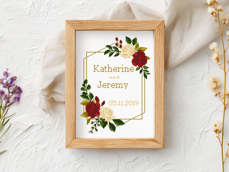May include: A framed cross-stitch embroidery featuring a gold geometric border, red and white roses, and green leaves. The text reads Katherine and Jeremy with the date 05.11.2019 in a gold font, all displayed within a light-colored wooden frame.