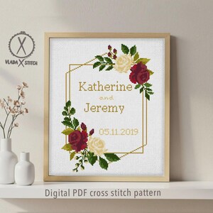 May include: A cross stitch pattern in a light wood frame featuring a gold geometric border, red and cream roses, green leaves, and the text Katherine and Jeremy 05.11.2019. The logo VLADA STITCH is visible in the upper left corner of the design.
