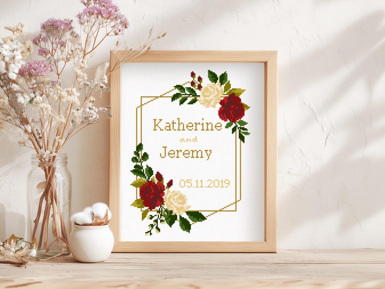 May include: A framed cross-stitch embroidery featuring a gold geometric border decorated with red and cream roses and green leaves. The text reads Katherine and Jeremy with the date 05.11.2019 in a gold-colored font on a white fabric background.