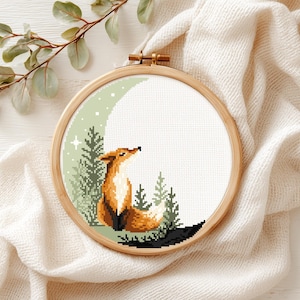 May include: A circular wooden embroidery hoop containing a cross-stitch pattern of an orange fox sitting in a forest of green pine trees against a large, pale green crescent moon background with small white stars scattered across the design.