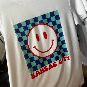May include: White t-shirt with a blue and green checkered design, a red smiley face, and the text "KANSAS CITY" in red.