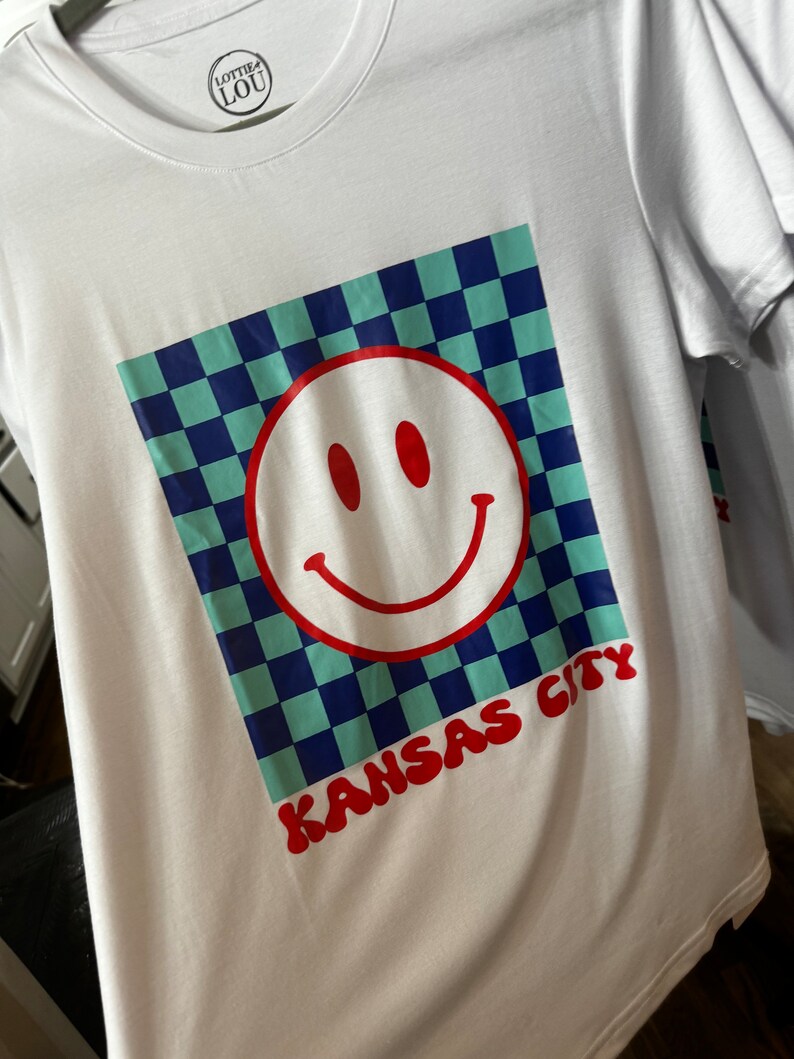May include: White t-shirt with a blue and green checkered design, a red smiley face, and the text "KANSAS CITY" in red.