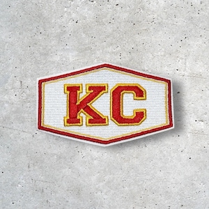 May include: A white, red, and gold patch with the letters "KC" in a bold, blocky font. The patch has a red border and a gold outline around the letters. The patch is in a unique hexagon shape.
