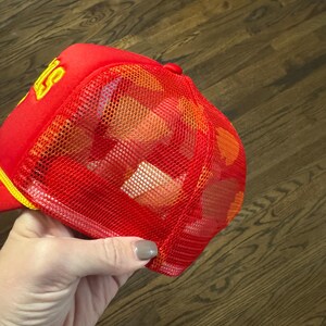 May include: Red and yellow mesh trucker hat with a yellow logo. The logo is a stylized letter "S" with a yellow outline.