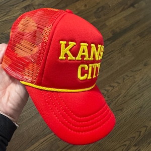 May include: Red and yellow trucker hat with the text "KANSAS CITY" embroidered on the front.