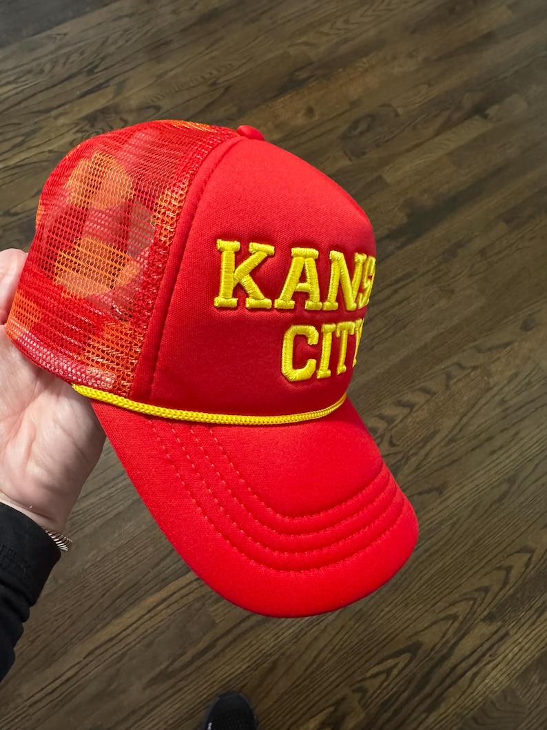 May include: Red and yellow trucker hat with the text "KANSAS CITY" embroidered on the front.