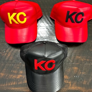 May include: Three trucker hats with a mesh back. Two hats are red with black "KC" lettering. One hat is black with red "KC" lettering.