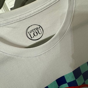 May include: White t-shirt with a black and white logo that says "Lottie Lou" in a circle. The shirt has a blue, green, and red checkered pattern at the bottom.