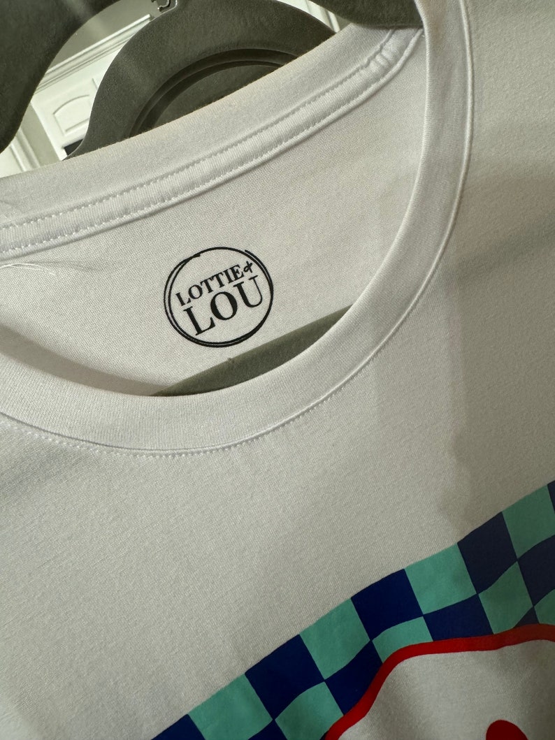May include: White t-shirt with a black and white logo that says "Lottie Lou" in a circle. The shirt has a blue, green, and red checkered pattern at the bottom.