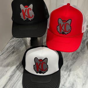 May include: Three trucker hats with a wolf head logo and the letters "KC" in red. The hats are black, red and white with black mesh backs.