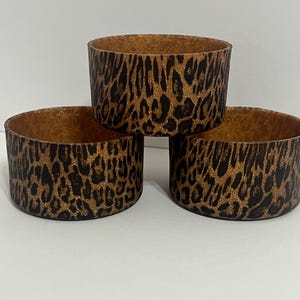Op de afbeelding: Drie cilindrische containers met een luipaardprint. De containers zijn bruin met zwarte vlekken en een glinsterend gouden interieur. De containers zijn gestapeld op een wit oppervlak.