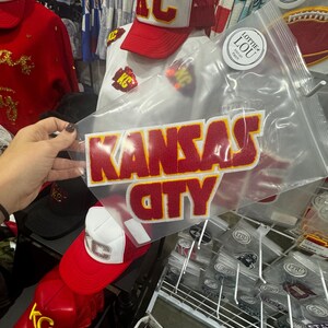 May include: A red and white trucker hat with the letters "KC" in gold embroidery. The hat is on a white t-shirt with a red iron-on patch that says "Kansas City".