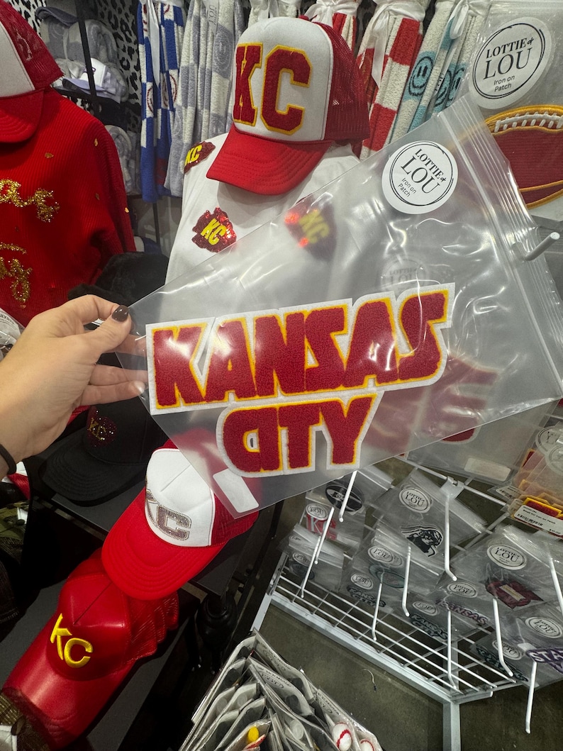 May include: A red and white trucker hat with the letters "KC" in gold embroidery. The hat is on a white t-shirt with a red iron-on patch that says "Kansas City".