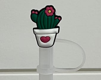 May include: A clear plastic straw with a cactus-shaped charm. The charm features a green cactus with a pink flower in a white pot with a red heart. The straw has a clear plastic loop attached.
