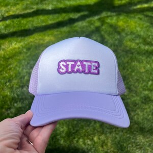 May include: A lavender trucker hat with a white mesh back. The hat has a purple patch with the word "STATE" in white letters.