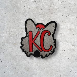 May include: A gray patch with a black border, featuring the red letters "KC". The patch has a stylized design, possibly representing an animal or mascot. The background is a textured, speckled surface.