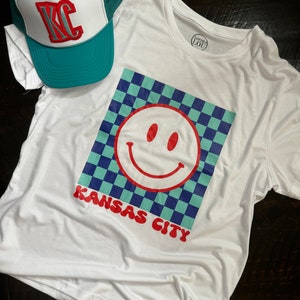 May include: White t-shirt with a blue and green checkered design and a red smiley face. The text "Kansas City" is printed in red below the design. A teal and white trucker hat with the letters "KC" in red is also in the image.