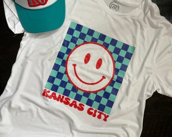 May include: White t-shirt with a blue and green checkered design and a red smiley face. The text "Kansas City" is printed in red below the design. A teal and white trucker hat with the letters "KC" in red is also in the image.