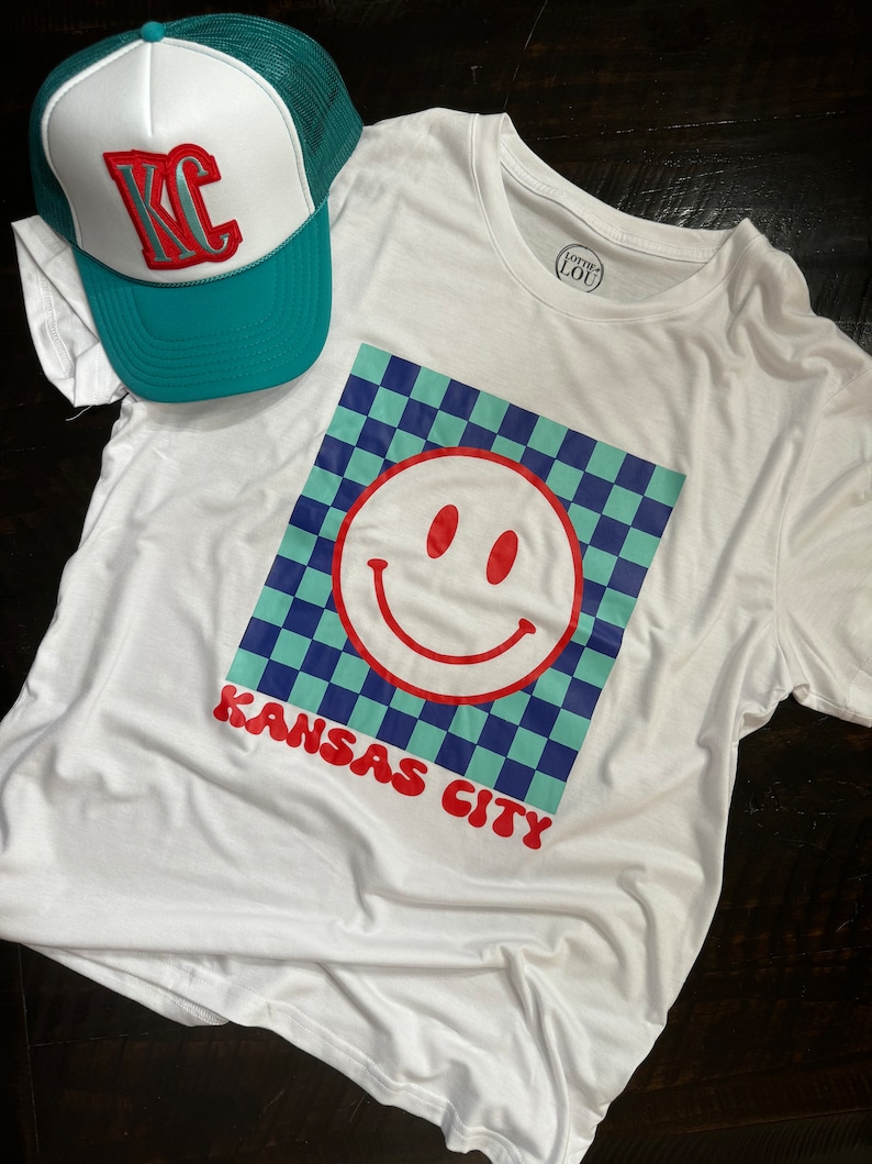 May include: White t-shirt with a blue and green checkered design and a red smiley face. The text "Kansas City" is printed in red below the design. A teal and white trucker hat with the letters "KC" in red is also in the image.