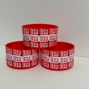 Puede incluir: Tres recipientes cilíndricos rojos apilados. Cada uno tiene una banda blanca con la palabra "USA" repetida, alternando con estrellas azules. Estos recipientes son probablemente para almacenamiento o exhibición.