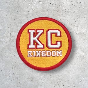 Op de afbeelding: Een ronde patch met een gele kern, rode rand en witte en rode letters. De letters "KC" staan bovenaan, met "KINGDOM" eronder. De patch is waarschijnlijk een geborduurd embleem.
