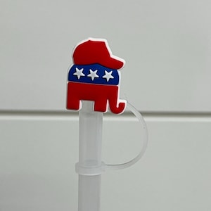 May include: A red, white, and blue elephant-shaped straw topper. The elephant is wearing a red hat and has three white stars on a blue background. The straw is clear plastic.