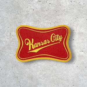 May include: Embroidered patch featuring the words "Kansas City" in yellow script on a red background. The patch has a yellow border and a unique, curved shape. The patch is likely for a jacket, bag, or other apparel.