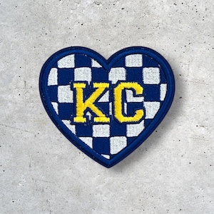 May include: Heart-shaped patch with a blue border and a checkered blue and white pattern. The letters "KC" are embroidered in yellow in the centre. The patch is likely for clothing or accessories.