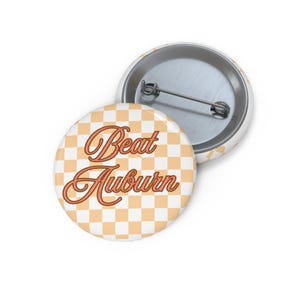 May include: Two round buttons. One button is face up, displaying the words "Beat Auburn" in a cursive font over a light orange and white checkered pattern. The other button is face down, showing the pin back.