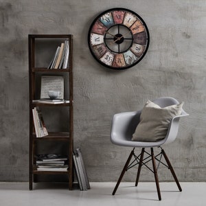 Könnte beinhalten: Ein grauer Sessel mit einem Kissen steht vor einem Holzregal mit Büchern und einer Tasse. Eine große, runde, mehrfarbige Uhr mit römischen Ziffern hängt an einer grauen Wand.