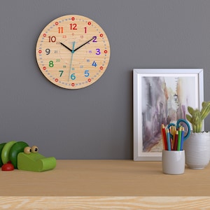Könnte beinhalten: Eine runde Wanduhr aus Holz mit bunten Zahlen und Zeigern. Das Zifferblatt hat ein helles Holzmaserungsmuster und Zahlen in verschiedenen Farben. Die Uhr ist an einer grauen Wand montiert, mit einem Holztisch im Vordergrund.